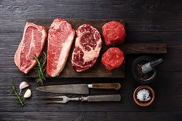 Fresho Software for fresh food wholesale suppliers and their customers - Meat Category - Quality slices of prime meat cuts laid out on a wooden cutting board with carving knife and fork, salt crystals and black pepper in a mortar and thistle